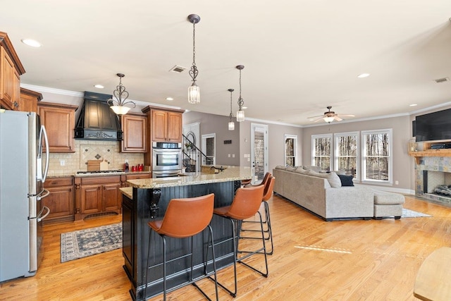 kitchen featuring light wood-style flooring, stainless steel appliances, custom exhaust hood, brown cabinets, and tasteful backsplash