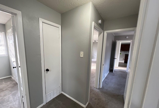 hallway featuring baseboards, dark carpet, a textured ceiling, and a textured wall