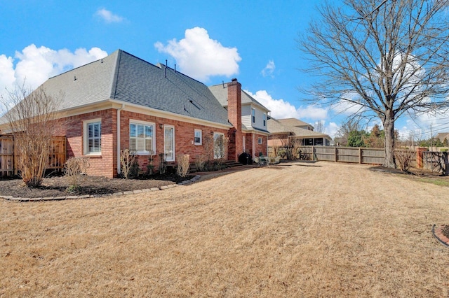 rear view of house with brick siding, a yard, a chimney, roof with shingles, and fence