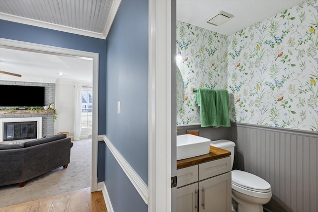 half bath featuring wallpapered walls, toilet, a wainscoted wall, wood finished floors, and a brick fireplace