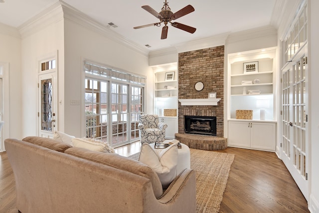 living area with wood finished floors, visible vents, a ceiling fan, ornamental molding, and a brick fireplace