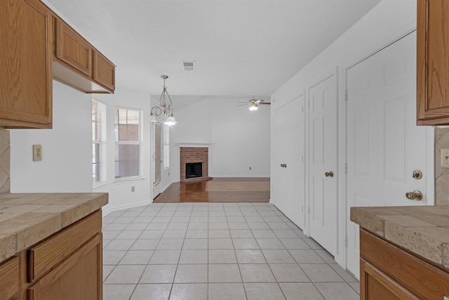 kitchen featuring tile counters, visible vents, a brick fireplace, light tile patterned flooring, and ceiling fan with notable chandelier