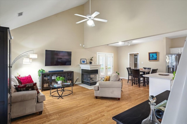 living area with light wood finished floors, visible vents, a multi sided fireplace, high vaulted ceiling, and baseboards