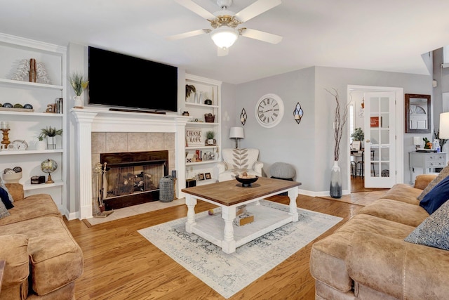 living room featuring baseboards, a tiled fireplace, built in features, light wood-type flooring, and a ceiling fan