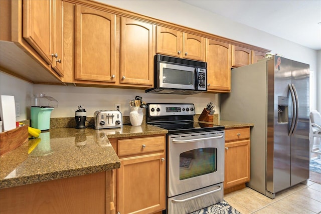 kitchen featuring light tile patterned floors, stainless steel appliances, dark stone countertops, and brown cabinets