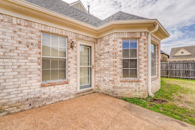 property entrance with brick siding, a patio area, roof with shingles, and fence