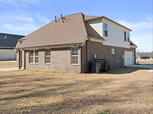 rear view of house with a lawn, roof with shingles, an attached garage, brick siding, and central AC unit