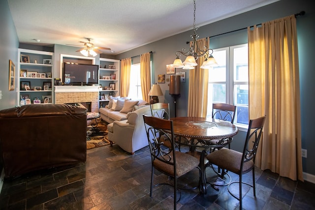 dining space with a brick fireplace, built in shelves, ceiling fan with notable chandelier, stone finish floor, and a textured ceiling