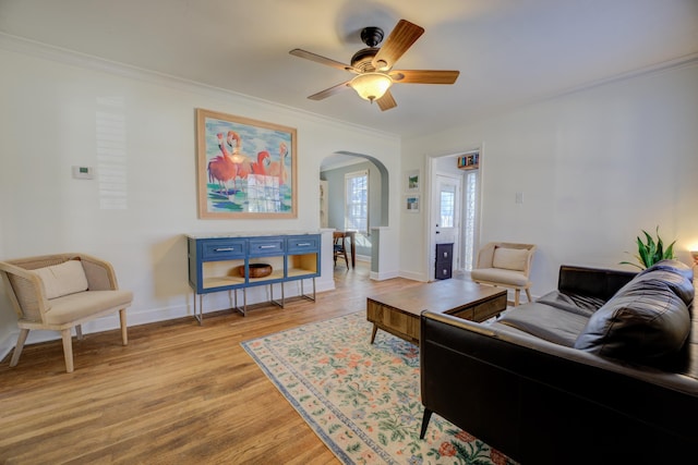 living area featuring light wood finished floors, baseboards, ornamental molding, arched walkways, and a ceiling fan