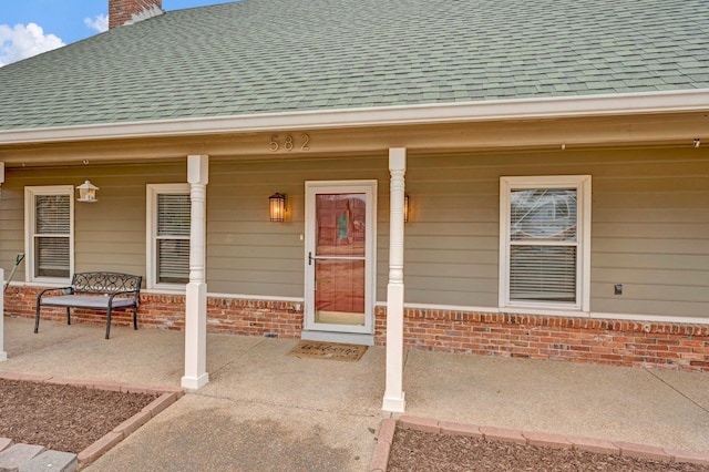 doorway to property with brick siding, a porch, and a shingled roof