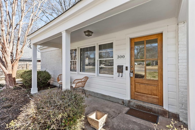 doorway to property featuring brick siding, covered porch, and fence