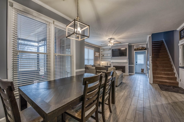 dining space featuring stairway, wood finished floors, a fireplace, ornamental molding, and ceiling fan with notable chandelier