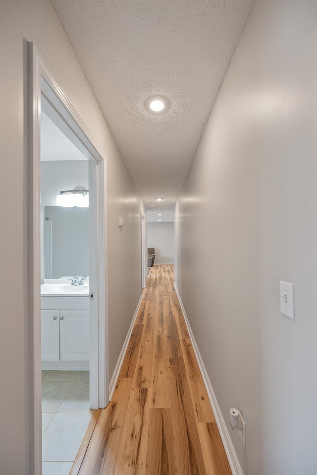 hall with a sink, baseboards, a textured ceiling, and light wood-style flooring