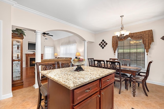 kitchen featuring ornamental molding, decorative columns, ceiling fan with notable chandelier, a warm lit fireplace, and arched walkways