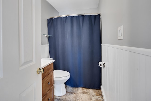 full bathroom with a wainscoted wall, toilet, a shower with shower curtain, stone finish floor, and vanity