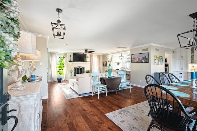 dining room featuring dark wood finished floors, a fireplace with raised hearth, ceiling fan with notable chandelier, and ornamental molding