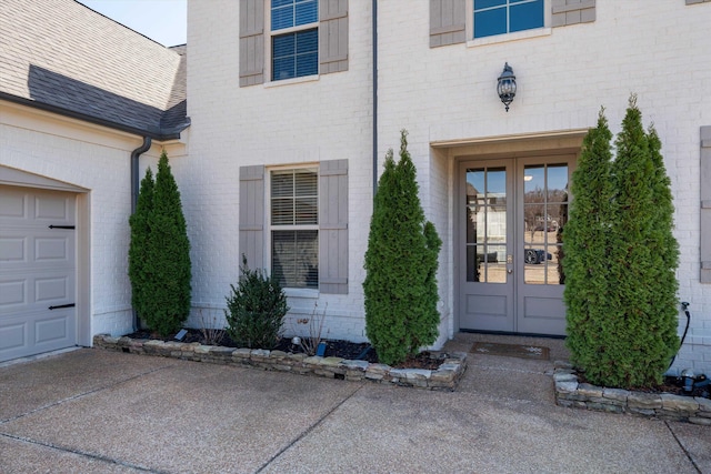 view of exterior entry with brick siding, french doors, a shingled roof, and driveway