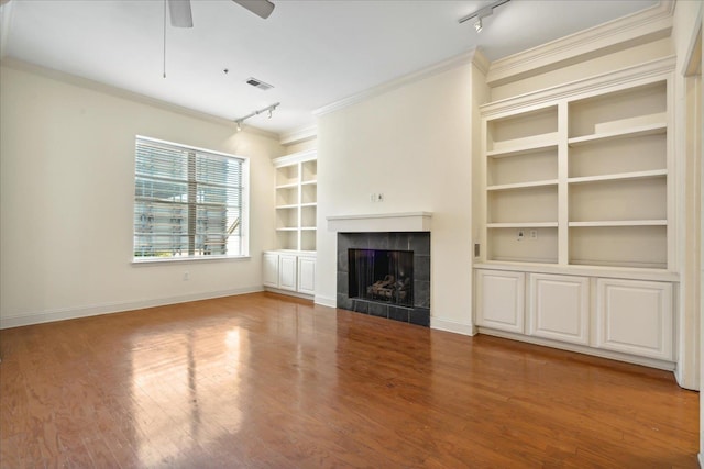 unfurnished living room featuring built in features, wood finished floors, a tile fireplace, ceiling fan, and crown molding