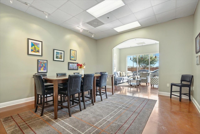 dining room with a drop ceiling, arched walkways, rail lighting, baseboards, and concrete flooring