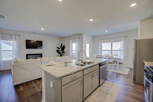 kitchen featuring light stone countertops, a center island with sink, a sink, dark wood-type flooring, and appliances with stainless steel finishes