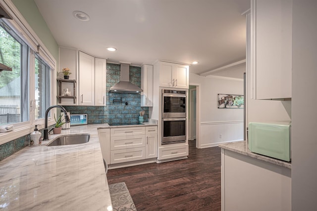 kitchen with light stone countertops, white microwave, stainless steel double oven, a sink, and wall chimney exhaust hood