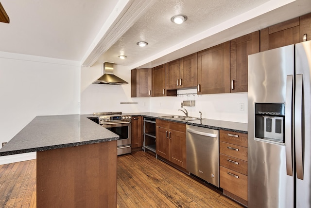 kitchen featuring dark wood finished floors, a peninsula, a sink, appliances with stainless steel finishes, and wall chimney exhaust hood