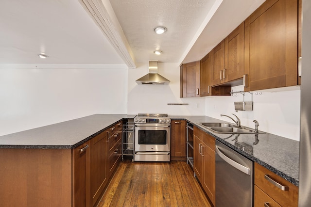 kitchen featuring brown cabinetry, a peninsula, a sink, appliances with stainless steel finishes, and wall chimney range hood