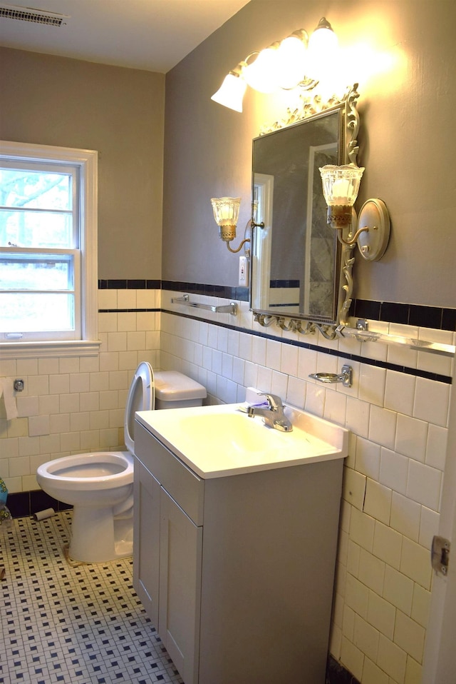 half bathroom featuring vanity, a wainscoted wall, visible vents, tile patterned flooring, and toilet