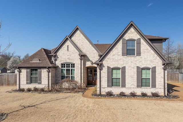 french country style house featuring brick siding, a shingled roof, and fence