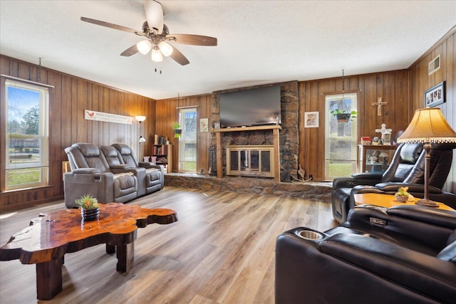 living room with a stone fireplace, wooden walls, a ceiling fan, and light wood-type flooring