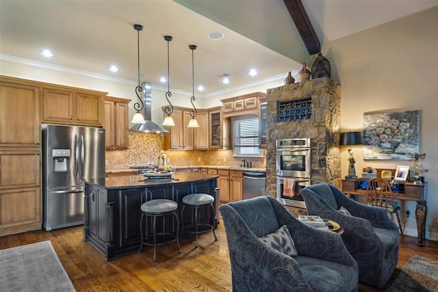 kitchen featuring wall chimney range hood, brown cabinets, decorative backsplash, an island with sink, and stainless steel appliances