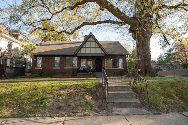 tudor-style house with brick siding, a chimney, a front yard, and roof with shingles