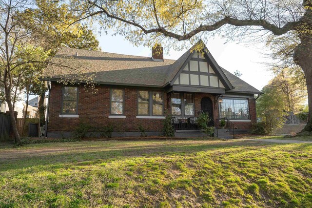 tudor-style house with brick siding, a chimney, a front lawn, and roof with shingles