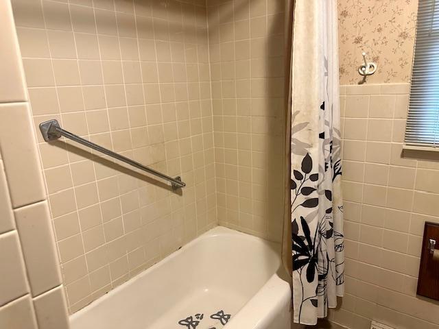 full bathroom with tile walls, wallpapered walls, shower / bath combo with shower curtain, and a wainscoted wall