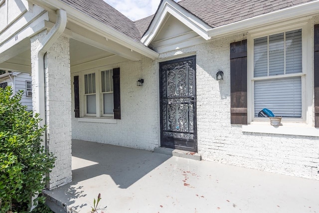 doorway to property featuring brick siding, a shingled roof, and covered porch