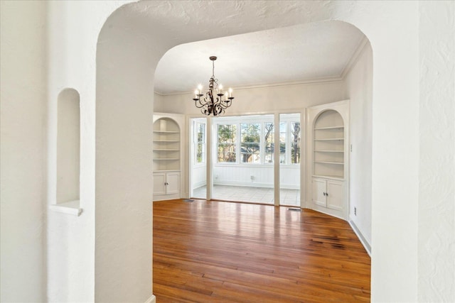 empty room featuring an inviting chandelier, built in features, ornamental molding, arched walkways, and wood finished floors
