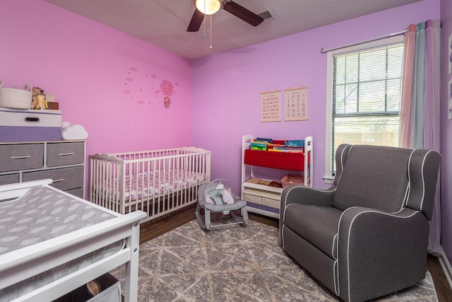 bedroom featuring visible vents, a nursery area, a ceiling fan, a textured ceiling, and wood finished floors