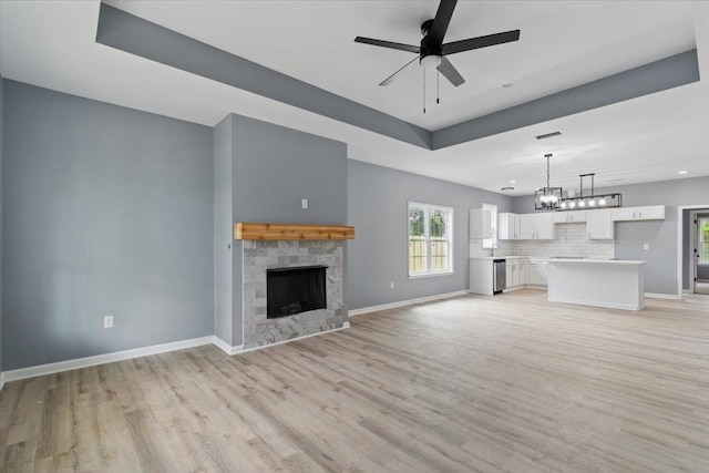 unfurnished living room featuring baseboards, a fireplace, a tray ceiling, ceiling fan with notable chandelier, and light wood-style flooring