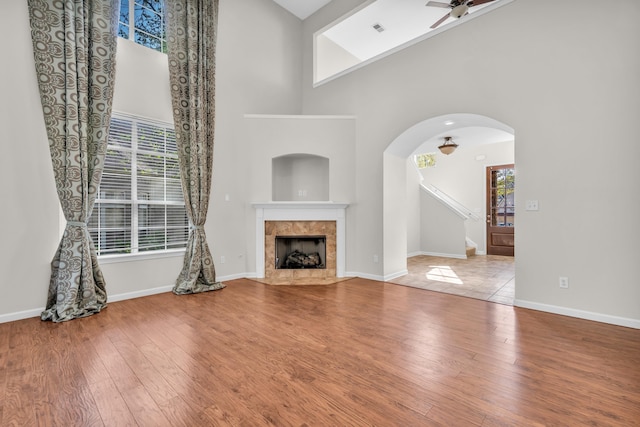 unfurnished living room with arched walkways, a high ceiling, ceiling fan, wood finished floors, and a tile fireplace