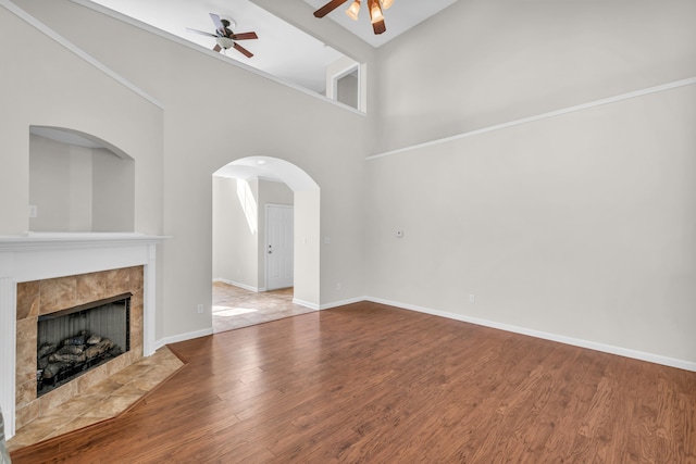 unfurnished living room with ceiling fan, arched walkways, wood finished floors, a tile fireplace, and baseboards