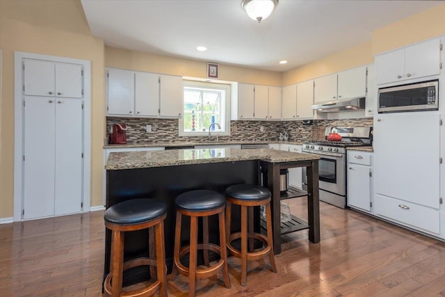 kitchen featuring a kitchen breakfast bar, under cabinet range hood, wood finished floors, stainless steel appliances, and backsplash