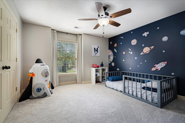carpeted bedroom with a textured ceiling, a crib, ceiling fan, baseboards, and visible vents