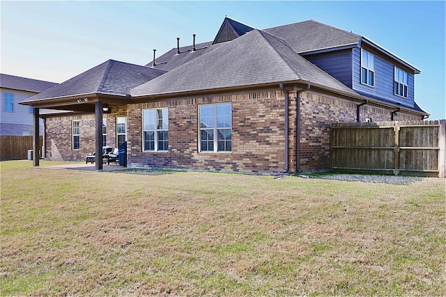 rear view of house with a lawn, fence, a patio, brick siding, and roof with shingles