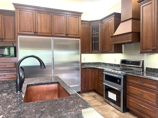 kitchen with appliances with stainless steel finishes, a sink, stone finish floor, premium range hood, and dark stone countertops