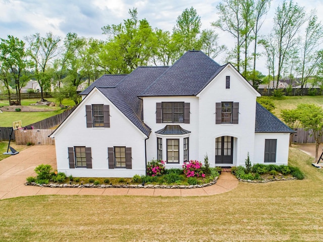 french provincial home with concrete driveway, a front lawn, roof with shingles, and fence