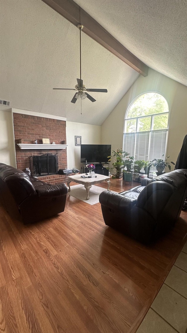 living area with a textured ceiling, visible vents, lofted ceiling with beams, a brick fireplace, and a ceiling fan