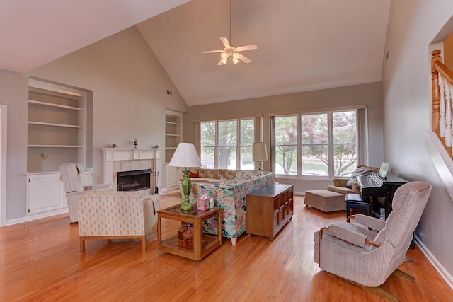 living room with a tiled fireplace, built in shelves, light wood finished floors, high vaulted ceiling, and a ceiling fan