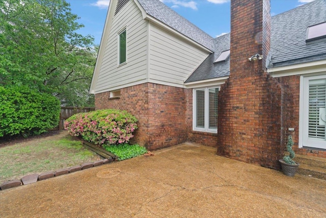view of side of home with a shingled roof, a patio area, a chimney, brick siding, and fence