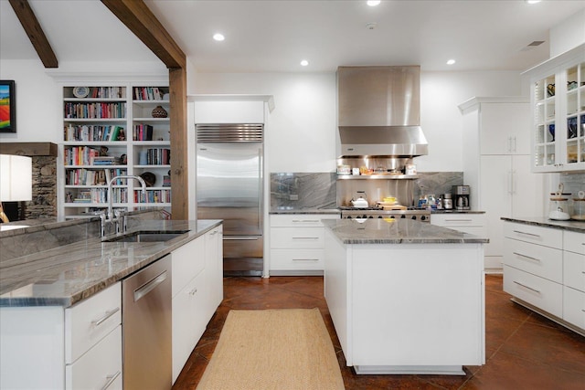 kitchen with a kitchen island with sink, light stone countertops, a sink, wall chimney exhaust hood, and appliances with stainless steel finishes