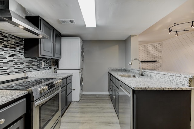 kitchen featuring wall chimney exhaust hood, stainless steel appliances, a peninsula, visible vents, and stacked washer and clothes dryer